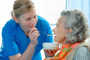 A female caregiver provides in-home assistance for an elderly woman by feeding her a healthy meal.