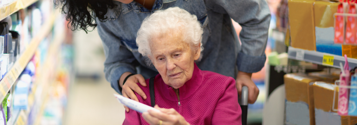 Caregiver pushing an older woman in a wheelchair through a grocery aisle as she reads a shopping list