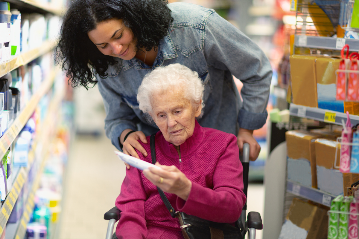 Caregiver pushing an older woman in a wheelchair through a grocery aisle as she reads a shopping list